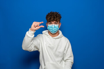 I need some more. Portrait of a young man in a surgical medical mask and a white sweatshirt making the gesture of a little more and begging for something. Indoor studio shot, isolated blue background.