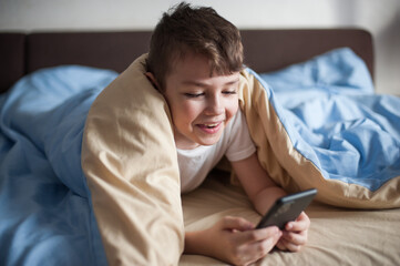 boy of European appearance lies under a blanket with a phone in his hands and communicates with a smile in an online chat