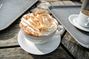 Closeup for a big white cup of hot chocolate with whipped cream and cocoa sprinkles on top, cup with drink standig on outdoors wooden table next to grey plastic trays.