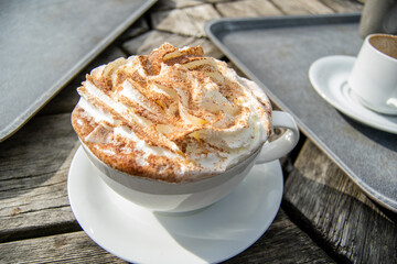 Closeup for a big white cup of hot chocolate with whipped cream and cocoa sprinkles on top, cup with drink standig on outdoors wooden table next to grey plastic trays.