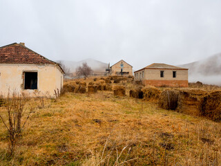 Large area with haystacks on dry grass and abandoned houses surrounded by thick cloudy fog. Countryside in the highlands.