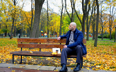 a young man sits on a bench in an autumn park and reads a book, holding a cup of coffee or tea in his other hand. education development concept
