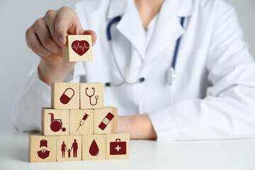 Doctor building pyramid of wooden cubes with different icons at white table, closeup. Insurance...