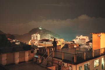 A night, long exposure shot of a residential favela area in Babilonia, in Leme district of Rio de Janeiro, Brazil, with multiple illuminated roofs of houses and hotels, and a mountain behind