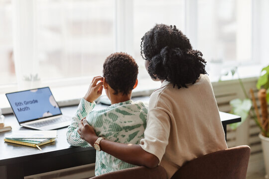Back view portrait of caring African American mother and daughter studying at home together and watching online lesson via laptop