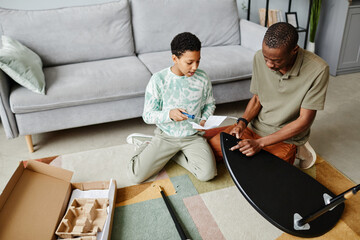 High angle portrait of teenage black girl helping father assembling furniture at home, copy space