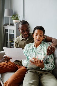 Vertical Portrait Of African American Man Helping Daughter Doing Homework While Sitting On Sofa At Home