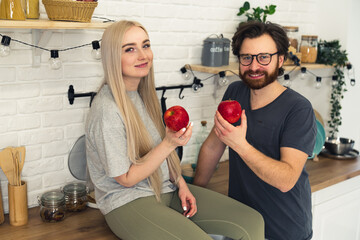 Obraz premium young married caucasian couple sharing healthy lifestyle, holding a red apple while being in the kitchen and looking at camera. High quality photo