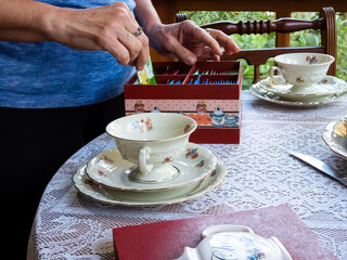 KPM porcelain tea set, decorared with flowers on a table. White lace tablecloth, silver knife, saucer, cup. A woman wearing blue is picking a tea bag on a red decorated tea box (hands only)