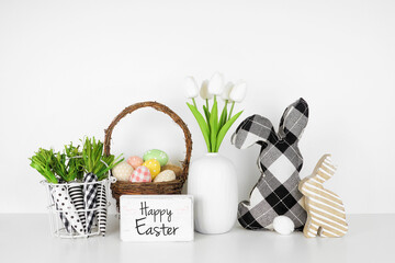 Happy Easter greeting with farmhouse style Easter decor and spring flowers on a white shelf against a white wall. Bunnies, basket of eggs, carrots and tulips.