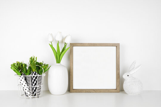 Mock up wood frame with Easter decor on a white shelf. Cloth carrots, knit bunny and tulip flowers. Square frame against a white wall. Copy space.