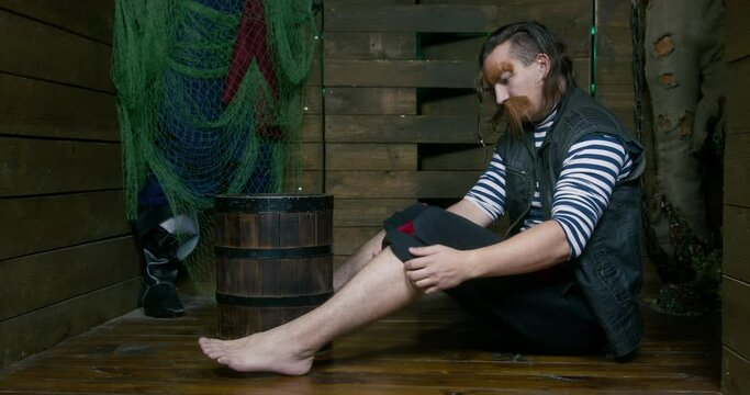 Barefoot pirate with long mustache rolls up black pants sitting near barrel ON wooden floor in brown cabin of pirates ship