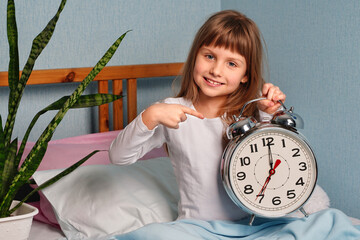 Huge alarm clock in the hands of a happy smile child girl in the bedroom. 7 o'clock in the morning.