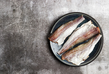 Raw Pollock (hake) fish fillet on a round plate on a dark gray background. Top view, flat lay