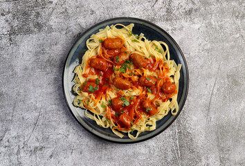 Tagliatelle pasta with sausages and tomato sauce on a round plate on a dark gray background. Top view, flat lay