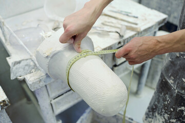 Hands of young worker of prosthetic factory holding measuring tape round plaster cast while working...