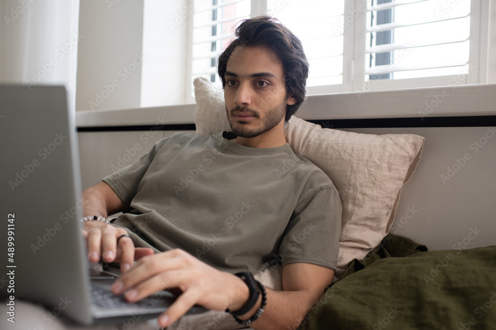 Poster young serious businessman typing on laptop keyboard while lying in bed and concentrating on network 