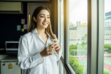 Young asian woman looking out window enjoying fresh new day feeling rested and drinking glass of milk at hotel.