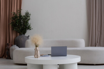 Composition of white round table with laptop and handmade vase against comfortable sofa and beige curtains by wall