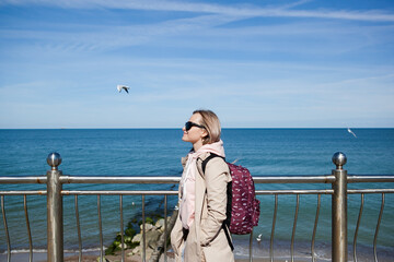 a blonde girl with a backpack on the background of a cold sea, seagulls in the sky. the concept of travel and trips