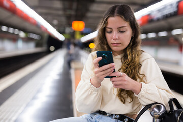Young european female watching mobile waiting the metro on the subway station platform