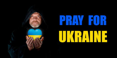 PRAY FOR UKRAINE.  Monk in a hood with a heart of Ukraine in his hands on a black background