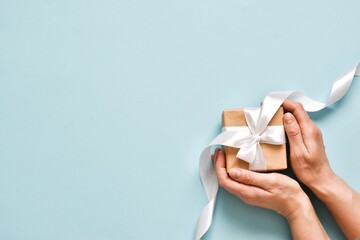 Female hands hold a gift box with a white ribbon on a blue background. The concept of Christmas, New Year, Valentine's Day and birthday. Top view with copy space