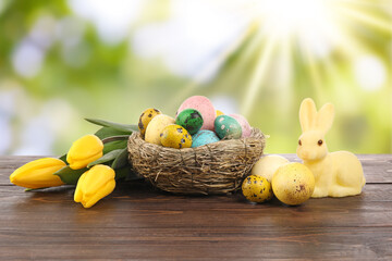 Nest with painted Easter eggs, tulip flowers and bunny on wooden table against blurred background