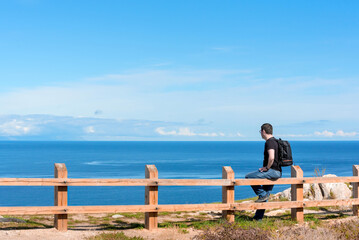 Fototapeta premium Man with backpack sitting on the fence at the end of earth watching on bright blue lake. Hiking or travel concept. Exploring the great outdoors. Beauty of nature