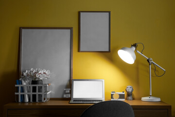 Modern workplace with laptop, organizer, frames and glowing lamp near yellow wall