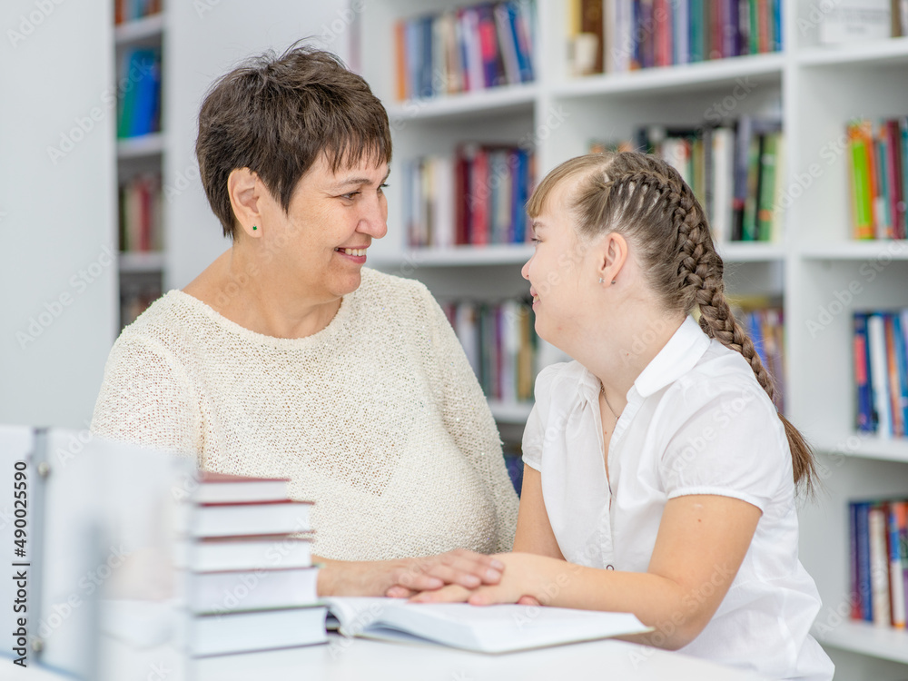 Wall mural Senior woman helps to girl with Downs syndrome doing homework at a school