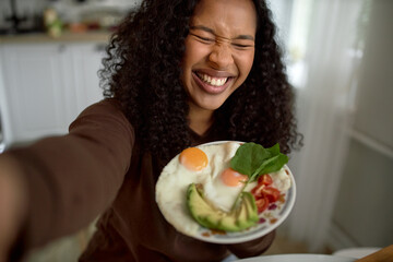 Laughing cheerful woman doing selfie with plate of yummy breakfast, squeezing or blinking eyes hard, having sweet toothy smile, standing against kitchen. Healthy food and lifestyle