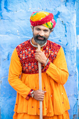 Happy traditional north indian man wearing colorful attire holding wood stick. Rajasthan male with turban and ethnic outfits. Culture and fashion. - obrazy, fototapety, plakaty