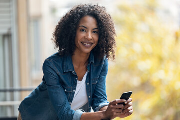 Cheerful woman using her smartphone while sending messages standing in the balcony at home.