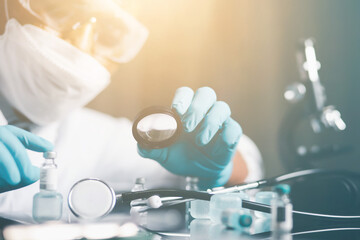 A young scientist in a white coat wearing safety goggles using a microscope zooms in on a sample and analyzes the virus.
