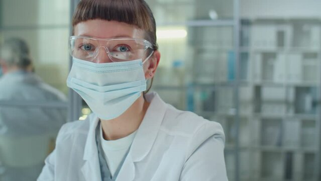 Portrait of young female scientist in protective mask and glasses sitting in laboratory and posing on camera
