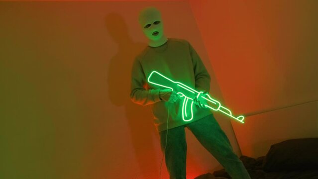 A gangster in a white mask holds a neon machine gun. A young guy is standing in an ultraviolet room on a bed. Video 4k for a rap clip in a neon room.