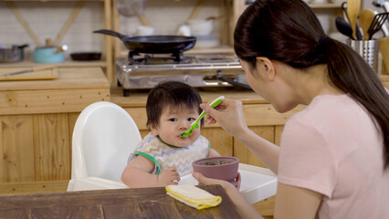 over the shoulder shot asian mom is stirring and scooping solid food in the bowl and feeding her...