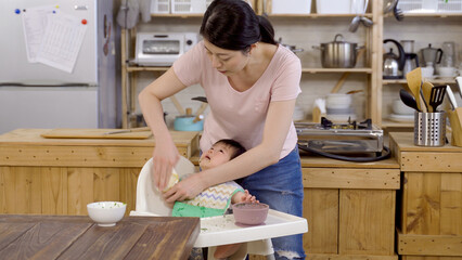 Korean mom standing and bending over is cleaning hands for her upset child on the high chair with cloth after meal at background modern home kitchen.