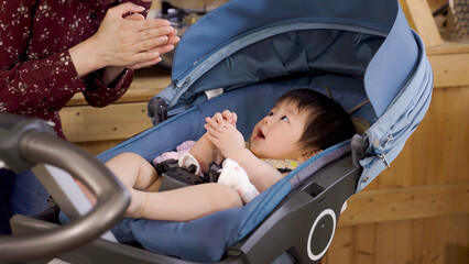 cropped shot innocent Korean baby lying in the stroller at home is looking at her mom who is interacting with her by singing and clapping hands.
