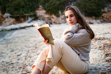 Relaxed woman sitting on the sand reading a book on a beach in spring clothes.