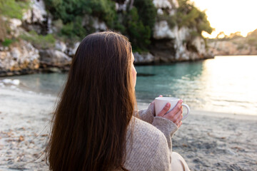 Unrecognizable woman drinking from a mug, sitting in the sand facing the sea, enjoying the sunset views of the natural landscape with spring clothes. Concept of enjoying coffee or tea.