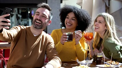 Video of bearded man taking selfie with female friends while having drink at terrace restaurant.