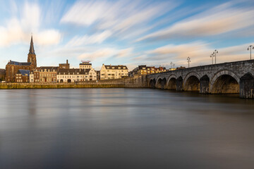 Obraz premium Maastricht, Netherlands 02-21-2022 Long exposure image creating moving clouds and silk water with a view on the Skyline of Maastricht and Wyck over the river Meuse and the Sint Servaas bridge