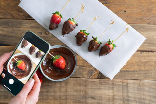 Strawberries with chocolate on a wooden background. Hand taking pictures with cell phone. Copy space.