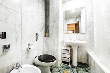 Bathroom clad in large white marble tiles with gray veining on the walls and green marble on the floor with white porcelain toilets and a shower with a curtain