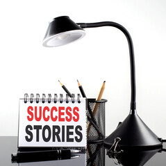 SUCCESS STORIES text on notebook with pen and table lamp on the black background
