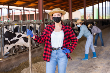 Obraz na płótnie Canvas Portrait of young positive woman farmer wearing face mask for disease and air pollution protection working in cowshed at dairy farm