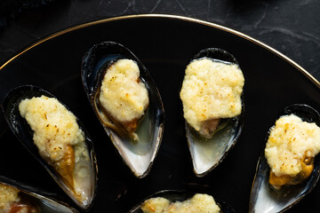 baked mussels in shells with creamy sauce poured with oil on a black plate on a dark background