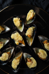 baked mussels in shells with creamy sauce poured with oil on a black plate on a dark background